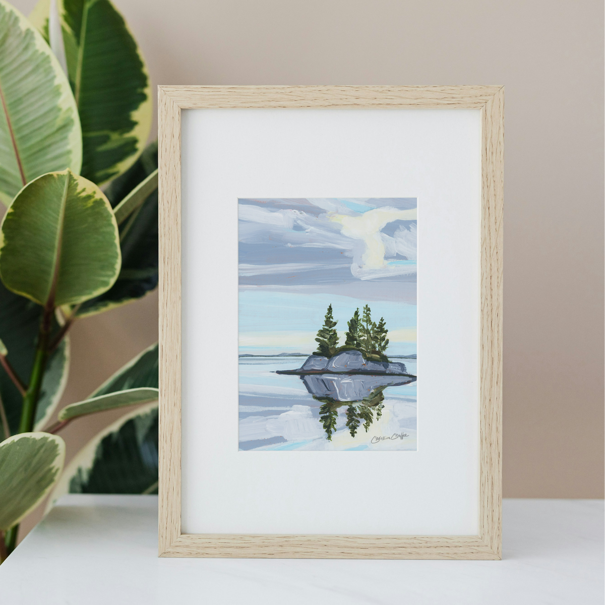 A framed art print depicting a serene landscape with a small island, pine trees, and a cloudy sky, styled on a white surface next to a green houseplant against a beige wall.